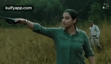 a woman in a green shirt is holding a gun in a field while a man holds a stick .