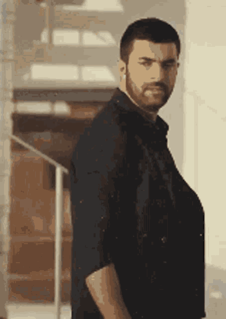 a man with a beard and a black shirt is standing in front of a staircase .