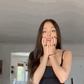 a woman in a black tank top is covering her mouth with her hands in a living room .