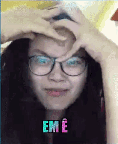 a woman wearing glasses is making a heart shape with her hands on her forehead with the word em e written on the bottom