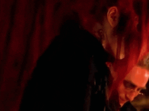 a woman with red hair and red lipstick is standing in a dark room with a man in the background .