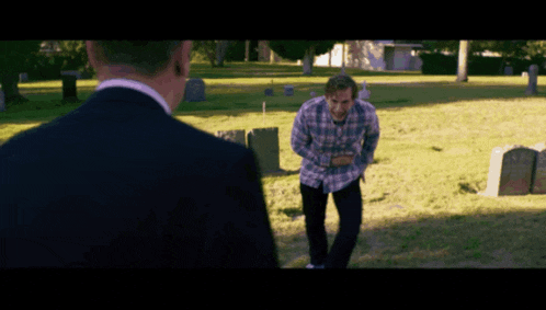 a man in a suit and plaid shirt is standing in a cemetery talking to another man