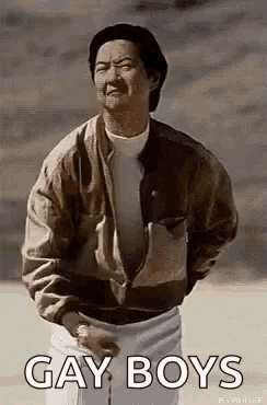 a man in a brown jacket and white shorts is standing in front of a wall and says `` gay boys '' .