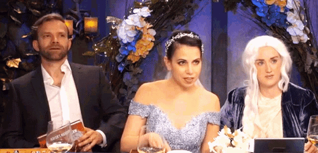 a man in a suit sits next to a woman in a blue dress and a woman in a white wig ..