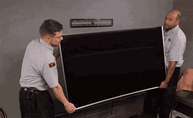 two men carrying a large flat screen tv with a sticker on their shirt that says ' sf ' on it
