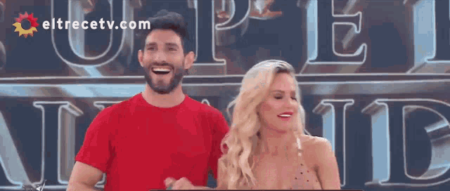 a man and a woman are laughing in front of a sign that says eltrecetv.com