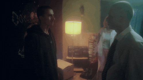 two men standing next to each other in a dark room with a lamp