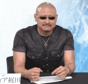 a man wearing sunglasses sits at a table in front of a sign that says mc