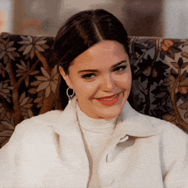a woman wearing a white jacket and earrings smiles while sitting on a chair