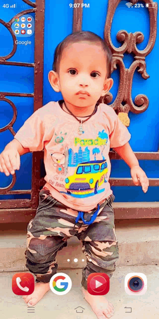 a baby is standing in front of a blue fence and a phone screen shows that it is 9:07 pm
