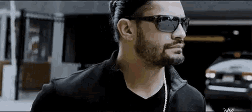 a man with a beard and sunglasses is wearing a black jacket and a black shirt .