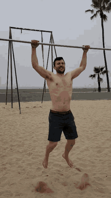 a shirtless man is hanging upside down on a bar on a beach