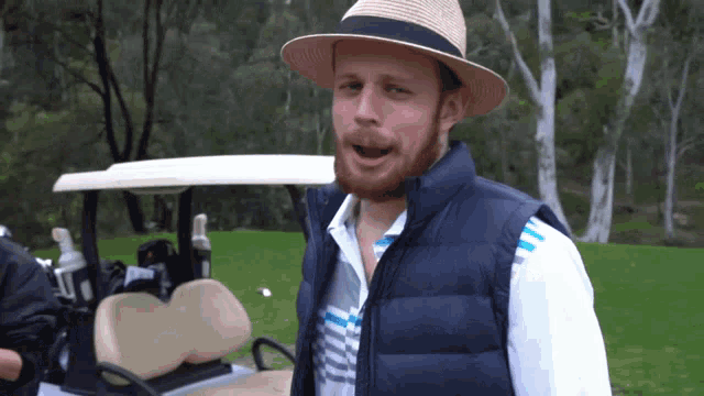 a man with a beard wearing a hat and a vest stands in front of a golf cart