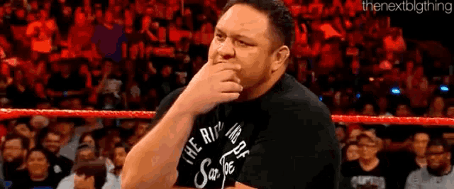 a man in a black shirt is standing in a wrestling ring with his hand to his mouth .