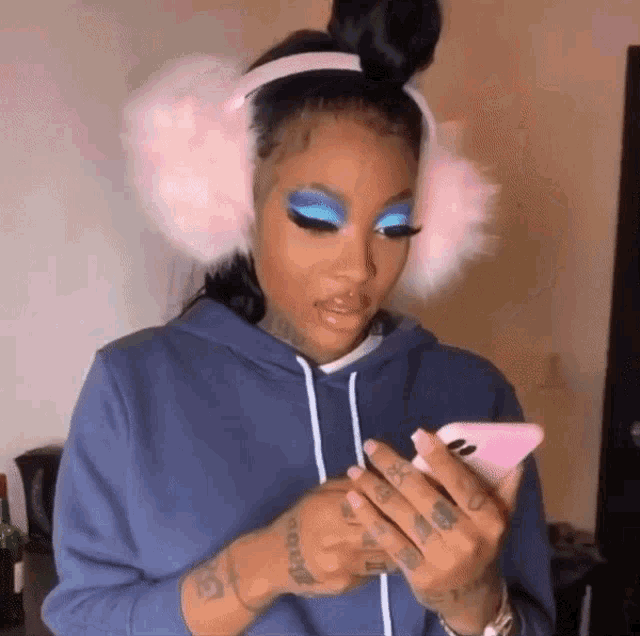 a woman wearing pink ear muffs and blue eye shadow looks at her phone