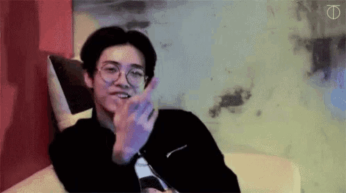 a young man wearing glasses is sitting on a couch and making a heart shape with his hand .
