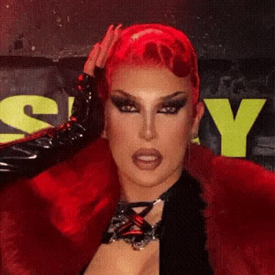 a drag queen with red hair and black gloves is holding her hair in front of a yellow sign .