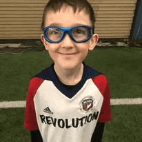 a young boy wearing glasses and a revolution jersey is smiling .