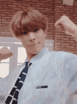 a young man in a blue shirt and tie flexes his muscles in front of a brick wall