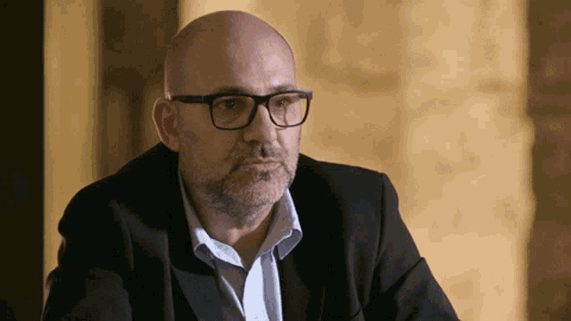 a bald man with glasses and a beard wears a black suit