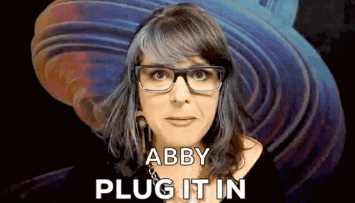 a woman wearing glasses is making a funny face and saying `` abby plug it in '' .