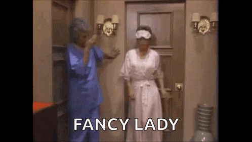 two women are standing next to each other in front of a door . one of the women is wearing a blindfold .
