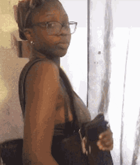 a woman wearing glasses is holding a purse and standing in front of a window .