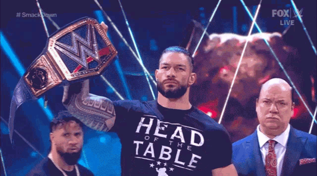 a man wearing a head of the table shirt holds a wrestling belt