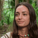 a close up of a woman 's face in the woods . she is wearing a necklace and smiling .