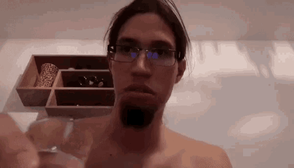 a shirtless man with glasses and a beard is holding a glass in his hand .
