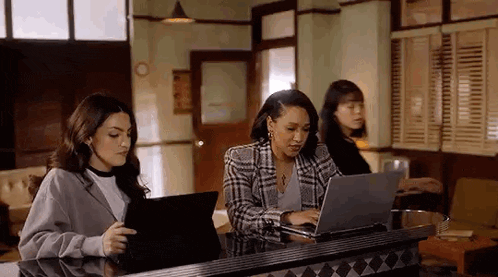 three women are sitting at a table using laptops and tablets .