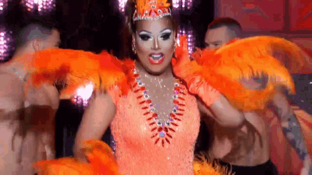 a drag queen is wearing orange feathers and a crown while dancing on stage .