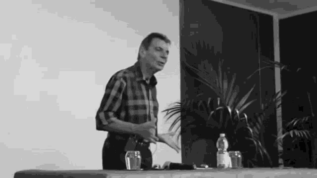 a man in a plaid shirt is standing in front of a table with a bottle of water and two glasses on it .
