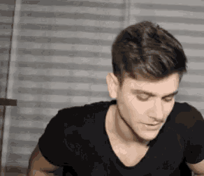 a man in a black t-shirt is sitting in front of a striped curtain and looking at the camera .