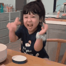 a little girl is sitting at a table with her arms in the air and making a funny face .