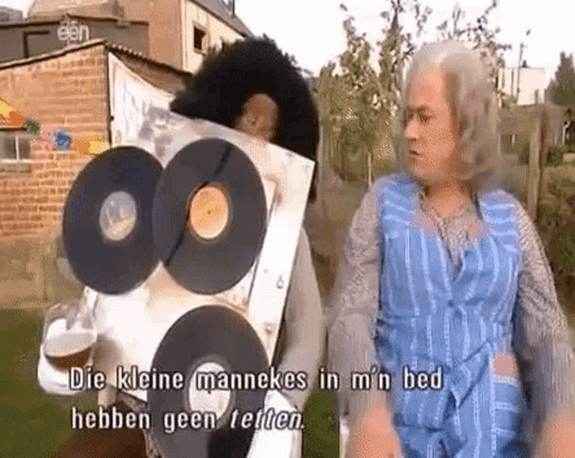 a man in a blue apron is holding a bunch of records while talking to another man .