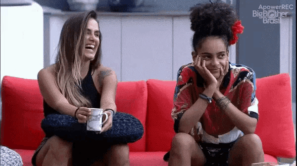 two women are sitting on a red couch and one is holding a mug