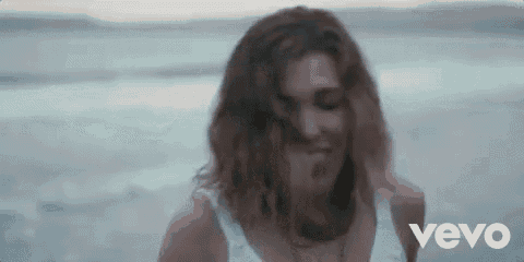 a woman in a white tank top is standing on a beach with the word vevo behind her