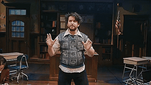 a man with a beard is standing in a classroom wearing a sweater vest and giving a thumbs up .