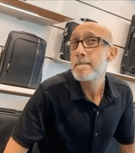 a bald man with glasses and a beard is sitting in front of a shelf with luggage .