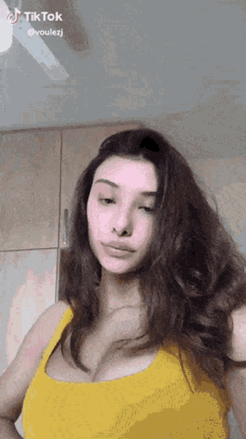 a woman in a yellow tank top is standing in front of a ceiling fan and looking at the camera .