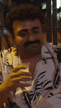 a man with a mustache is drinking from a glass while wearing a floral shirt .