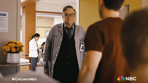a man in a lab coat stands in a hallway with a nbc logo in the corner