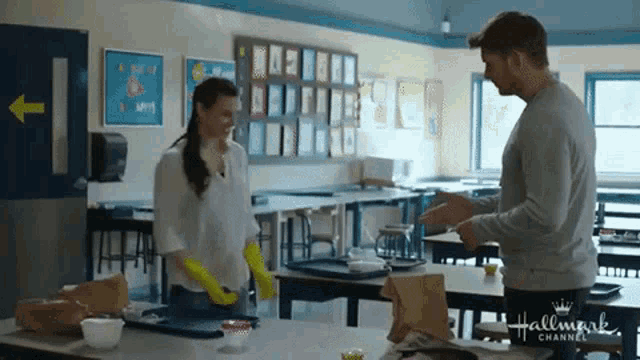 a man and a woman are standing in a classroom . the woman is wearing yellow gloves .
