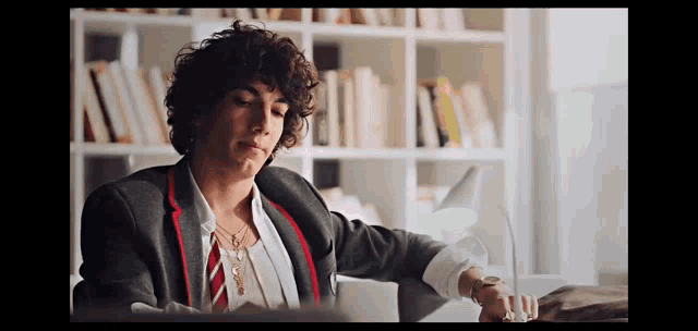a woman with curly hair is sitting at a desk in front of a bookshelf and looking at her watch .