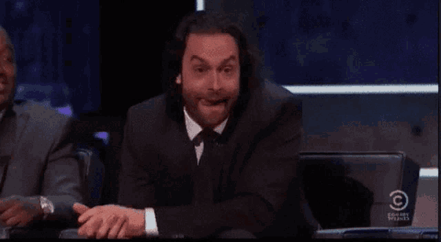 a man in a suit and tie is sitting in front of a screen that says c on it