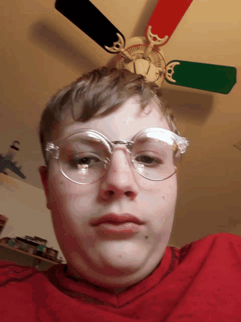 a young man wearing glasses looks at the camera with a ceiling fan in the background