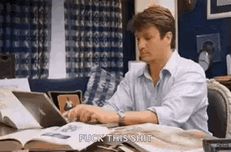 a man is sitting at a desk using a laptop computer while looking at a book .
