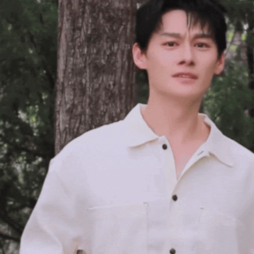 a young man wearing a white shirt is standing in front of a tree