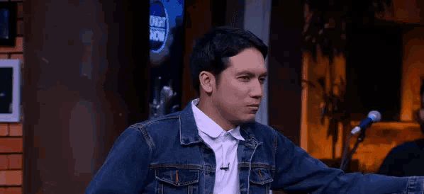 a man in a denim jacket is sitting in front of a microphone and says hi sayanggg
